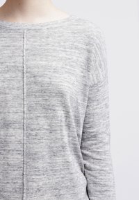 Lichtgrijze gebreide trui met een zachte textuur, boothals en subtiel verticale naad details. De stof heeft een gemêleerd patroon en een ontspannen pasvorm.