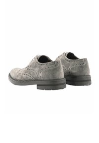 Scarpe brogue in suede grigio con una finitura liscia, perforazioni decorative e una robusta suola in gomma nera. La vista posteriore mostra la punta arrotondata e i lacci.