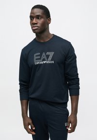 Muž v tmavě modré mikině a kalhotách EA7 Emporio Armani stojí na světlém neutrálním pozadí a dívá se vlevo.