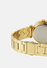 Kultakello, jossa on metallinen rannetaitto ja chevron-kuvio. Pyöreä kotelo, jossa on sileä, heijastava pinta ja painikkeella toimiva lukko.