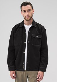 Veste en denim noir avec surpiqûres blanches contrastantes, fermeture à boutons sur le devant, une poche poitrine et deux poches latérales. Col classique avec manches longues.