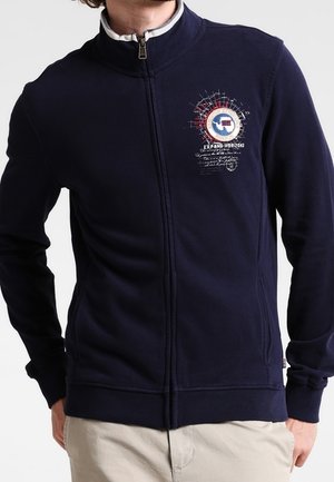 Veste zippée marine en tissu doux avec un col haut. Présente un design graphique sur la poitrine, des poignets côtelés et un ourlet pour un look ajusté.