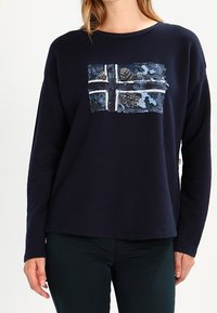 Mujer vistiendo una camiseta de manga larga en azul marino con un diseño estilizado de cruz nórdica en azul y blanco en el pecho, combinada con pantalones oscuros.