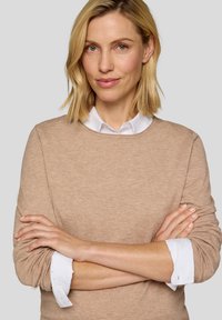 Hellbrauner Pullover mit Rundhalsausschnitt, der über einem weißen Hemd mit Kragen getragen wird. Der Stoff wirkt weich und hat ein einfaches, klares Design.