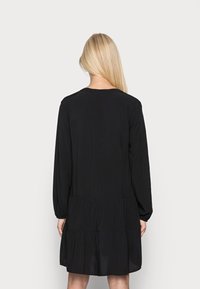 Vestido negro de manga larga con dobladillo en capas, corte holgado, escote redondeado y textura suave. Diseño sencillo sin adornos visibles.