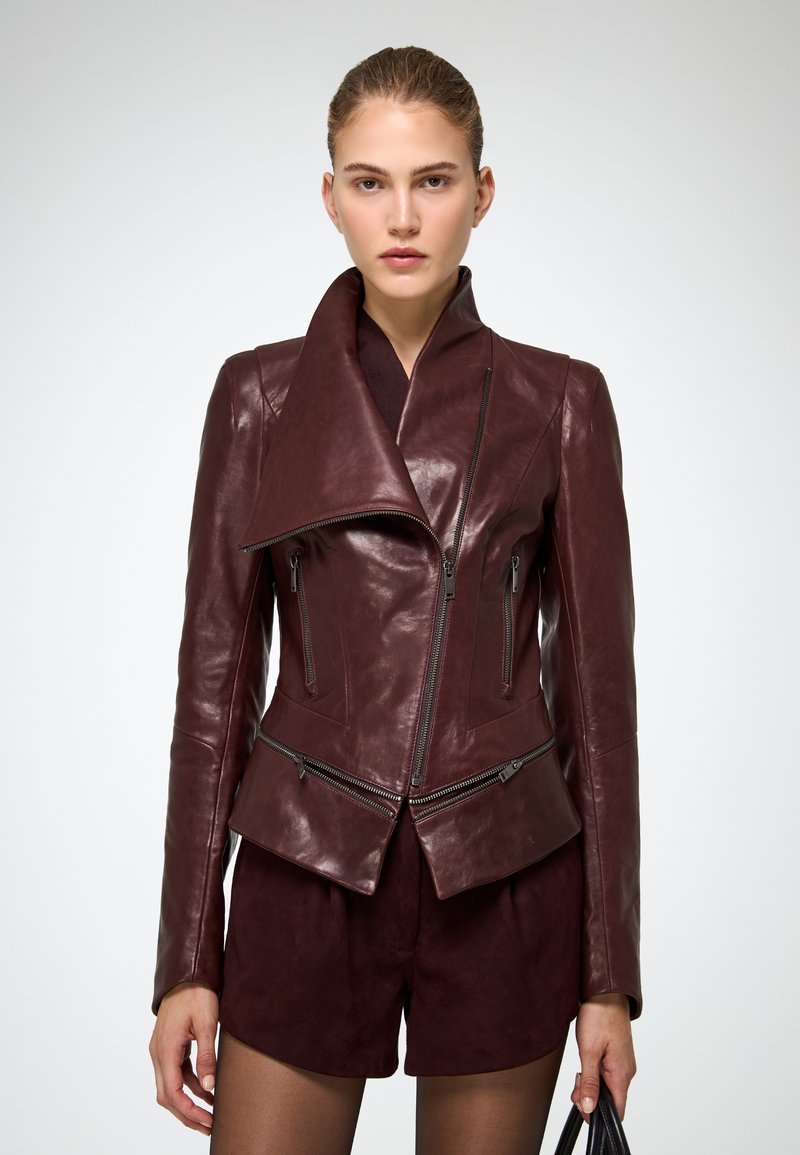 Burgundia nahast mootorrattajope, millel on asümmeetriline lukk, lai krae ja kitsad varrukad. Sile tekstuur koos eristuvate õmblustega.