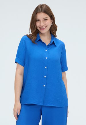 Donna sorridente con capelli castani che indossa una camicia azzurra a maniche corte con bottoni e pantaloni abbinati su uno sfondo chiaro uniforme.