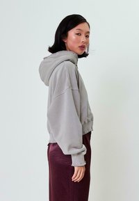 Geknipte grijze hoodie met geribbelde zoom en boorden. Zachte textuur, oversized pasvorm en een capuchon. Model draagt het met een bordeauxrode rok.