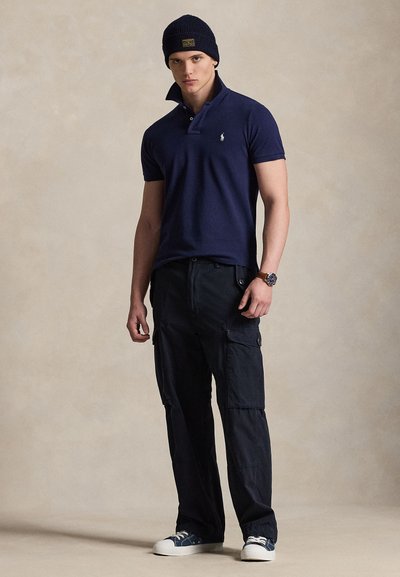 Joven con gorro azul marino, camiseta polo azul marino, pantalones cargo negros, zapatillas azules y blancas y reloj de pulsera, de pie frente a un fondo beige.
