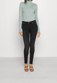 Un haut à col roulé côtelé vert clair associé à un jean skinny noir taille haute et des sandales noires à lanières. Design simple et ajusté.