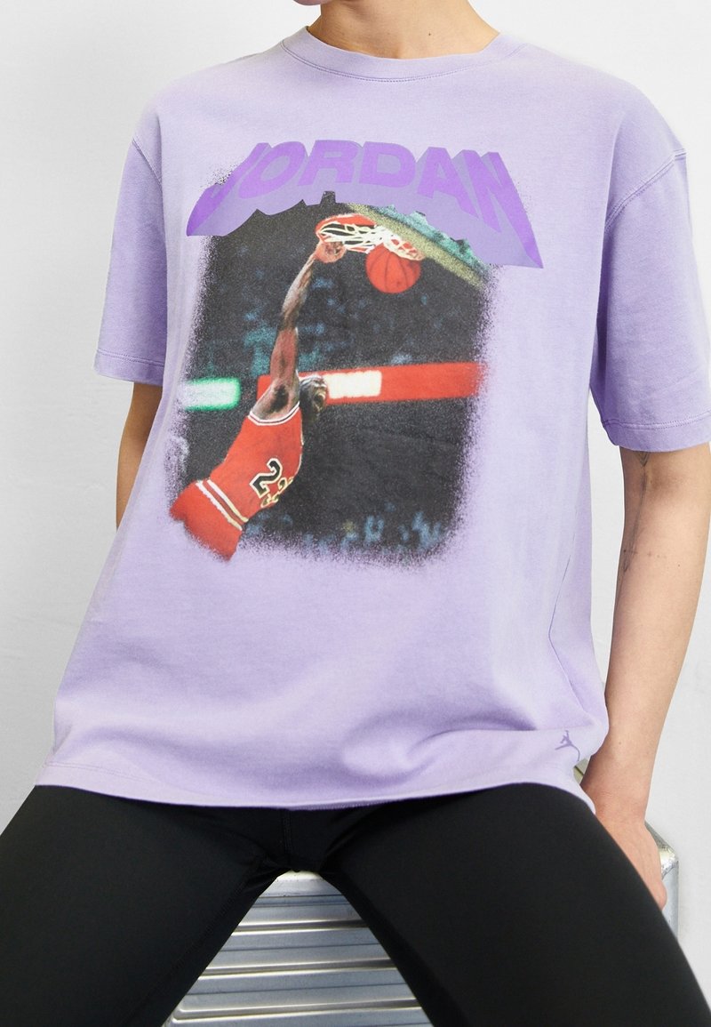 T-shirt en coton violet avec un graphisme représentant un joueur de basket-ball en maillot rouge, avec le texte "JORDAN" en lettres majuscules violettes au-dessus de l'image.