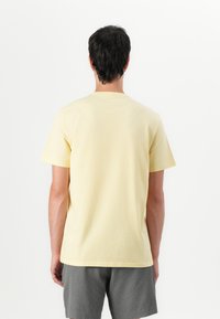 Camiseta de algodón amarilla con un cuello redondo clásico y mangas cortas, mostrada desde la parte de atrás. La tela parece suave y lisa.