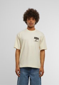 Cremefarvet t-shirt med korte ærmer, prydet med et mørkebrun "BEWARE" grafik på venstre bryst. Blødt bomateriale, afslappet pasform.