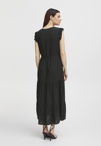 Gaun maxi hitam dengan desain bertingkat, lengan puff, dan pinggang elastis. Kainnya tampak halus dan ringan. Dipadukan dengan sepatu bertumit tinggi.