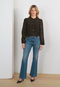 Black long-sleeved blouse with button closure and textured fabric, paired with blue flared jeans and black pointed-toe heels.