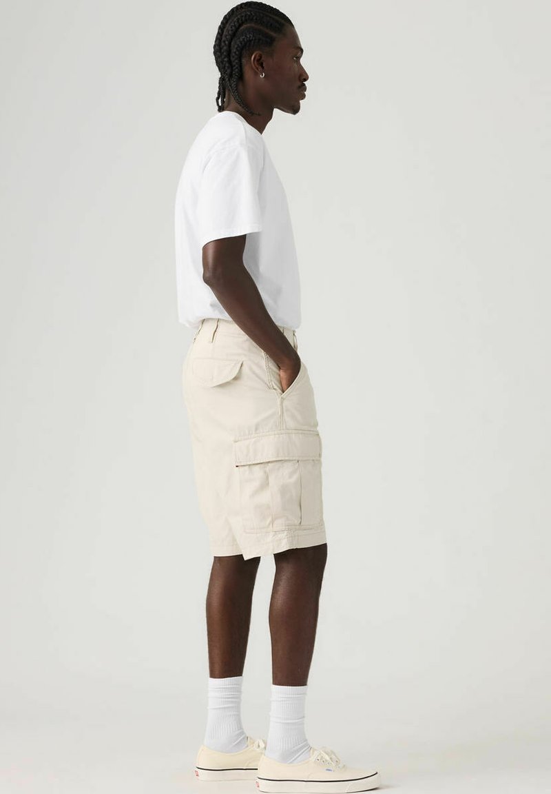 Shorts cargo beige avec multiples poches, portés avec un t-shirt blanc et des baskets crème. Le mannequin est de profil, portant des chaussettes blanches.