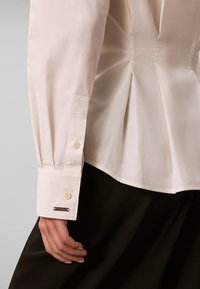 Gros plan d'une personne portant un chemisier crème à manches longues avec des poignets boutonnés et un pantalon noir, montrant la main et les plis latéraux du chemisier.