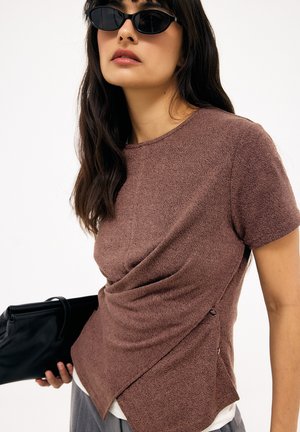 Vrouw met donker haar die zwarte cat-eye zonnebrillen draagt, een bruin textuur topje met korte mouwen en een zwarte clutch vasthoudt tegen een witte achtergrond.