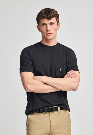 Zwarte crewneck t-shirt met korte mouwen en een borstzak; heeft een klein geborduurd logo; gedragen met beige broek en een zwarte gevlochten riem.