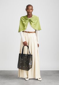 Γυναίκα που φοράει ελαφρύ πράσινο κασκόλ, λευκή μπλούζα με μακρύ μανίκι, κρέμα φαρδιά παντελόνια, λευκά παπούτσια, κρατώντας μια μαύρη πλέγμα αθλητική τσάντα με λουλούδια κεντημένα.