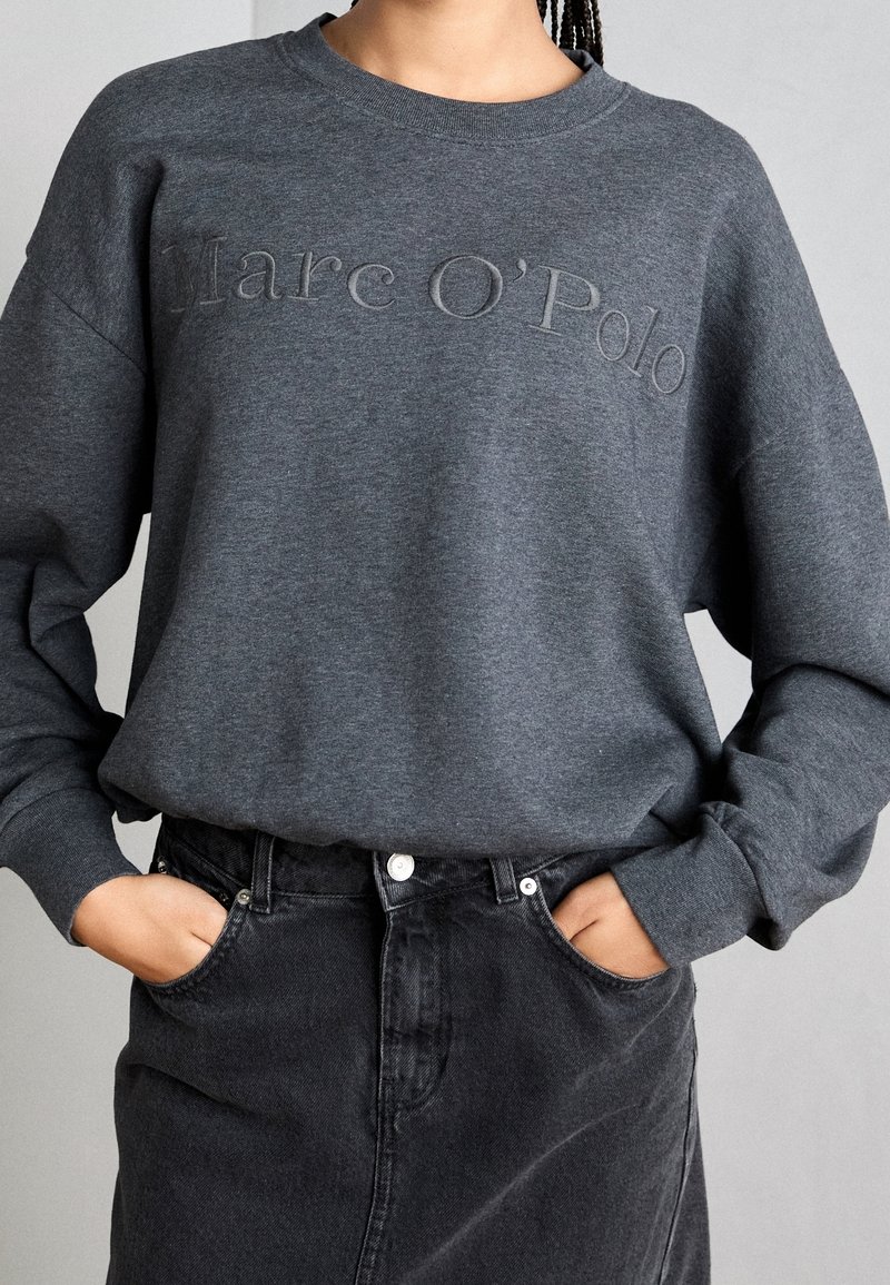 Personne portant un sweat-shirt gris foncé Marc O’Polo rentré dans une jupe en jean noire, les mains dans les poches.