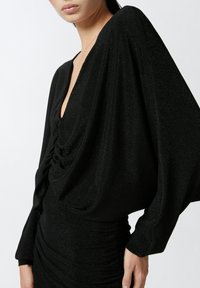 Abito nero con texture, scollatura a V profonda e maniche voluminose. I dettagli arricciati in vita creano una silhouette aderente.
