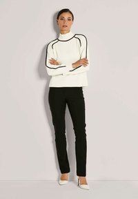 Frau, die mit verschränkten Armen vor einem einfarbigen Hintergrund steht, trägt einen weißen Rollkragenpullover mit schwarzer Paspelierung, schwarze Hosen und weiße High Heels.