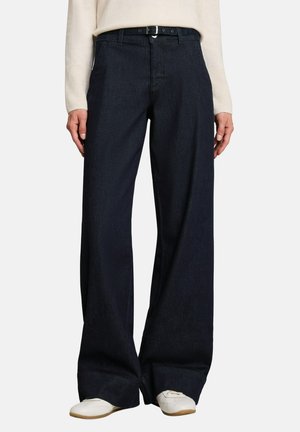 Persoon draagt donkere wijde jeans met een zwarte riem, een lichtkleurige top met lange mouwen en witte schoenen tegen een effen achtergrond.