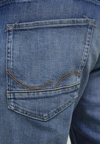 Jean en denim bleu avec une poche arrière dotée de coutures beiges et d'un design légèrement courbé. Le tissu a une texture douce et une finition délavée.