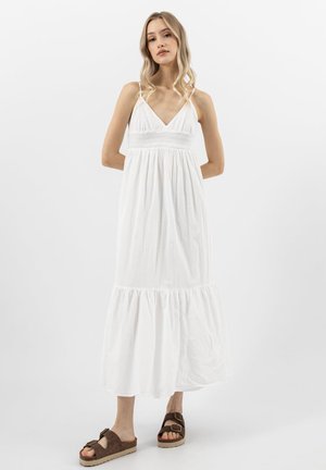 Žena stojí se založenýma rukama za zády, oblečená v bílé maxišatech bez rukávů a hnědých nazouvacích sandálech, na jednobarevném pozadí.