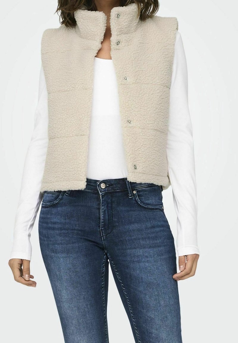 Femme portant un gilet polaire couleur crème par-dessus un haut blanc à manches longues et un jean denim bleu foncé, visible du cou aux hanches.