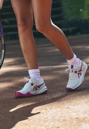 Primer plano de piernas con zapatillas de tenis blancas y rosas pisando una pista de tenis de tierra batida, sosteniendo una raqueta en el lado izquierdo.