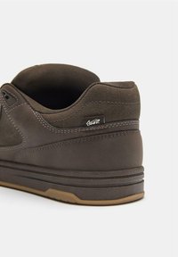 Hnedá kožená teniska s textilnými doplnkami, dizajn s nízkym strihom, kontrastné prešívanie a gumová podrážka. Má na boku etiketu s logom.