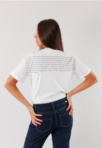 T-shirt bianco a maniche corte con testo stampato sulla schiena, abbinato a jeans di denim blu scuro. Modello di spalle, che mostra un fit casual.