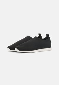 Svarte slip-on sko laget av pustende strikkemateriale, med en lett hvit såle og drag detaljer på hælen for enkel påføring.