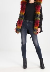 Svart jacka med flerfärgad fuskpälsdetalj, bar över en svart topp. Slim-fit mörkblå jeans och svarta ankelstövlar kompletterar outfiten.