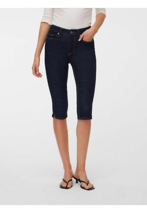 Femme portant un short en jean bleu foncé jusqu'aux genoux, un haut blanc et des sandales à talons noires, debout avec une main dans la poche.