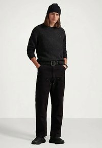 Άνδρας με μαύρη πλεκτή σκούφα, μαύρο πουλόβερ, μαύρο παντελόνι με ζώνη, μαύρα παπούτσια, όρθιος με τα χέρια στις τσέπες μπροστά σε απλή φόντο.