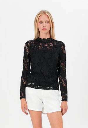 Top en dentelle noire à manches longues avec motifs floraux, col montant et manches transparentes. Associé à un short blanc.
