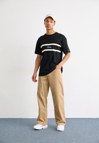 Juodas trumpas rankoves t-shirt su baltais horizontaliais dryžiais ir logotipu, derinamas su šviesiais plačiais velveto kelniais ir baltais sportbačiais. Šviesus kepurė.