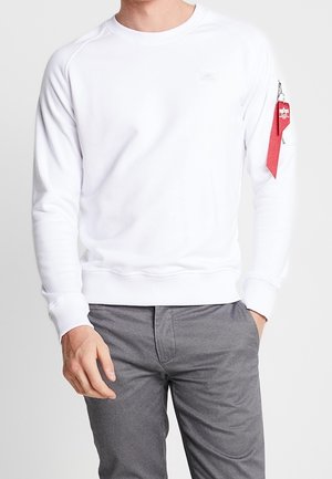 Homme portant un sweat-shirt blanc uni à manches longues avec une poche zippée rouge sur la manche gauche et un pantalon gris.