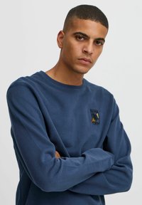 Marineblå hettegenser laget av bomull med lange ermer, rund hals og brodert «Born Duck»-grafikk på forsiden. Glatt tekstur.