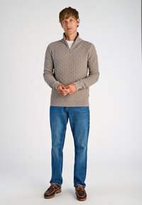 Beige gebreide trui met een ruitpatroon, half-rits kraag en ribbelmanchetten. Gepaard met blauwe jeans en bruine veterschoenen.