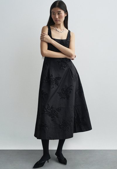Robe maxi noire avec un décolleté carré, des motifs floraux brodés et des détails ornés de perles ; associée à des collants noirs et des chaussures à bout pointu.