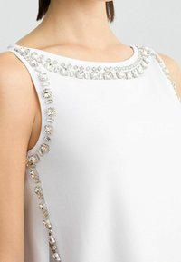 Top bianco senza maniche con scollatura tonda decorata con strass trasparenti lungo le spalle e le aperture delle braccia, che mettono in risalto accenti scintillanti.