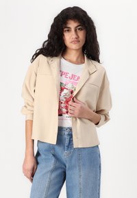 Beige button-up shirt with a collar, featuring two front pockets, worn over a colorful graphic t-shirt and paired with light blue jeans.