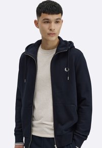 Granatowa bluza na suwak wykonana z miękkiego materiału, z kapturem ze sznurkiem, przednimi kieszeniami oraz białym haftowanym logo na piersi. Pod spodem jasnoszara koszulka.