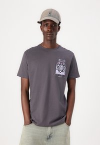 Grå bomullströja med en framsida som har en grafisk design med solmotiv. Kombinerad med en ljusbeige keps. Modellen står med händerna i fickorna.