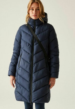 Mujer con abrigo de plumón acolchado largo azul marino con capucha y correa cruzada negra, de pie frente a un fondo claro y liso.