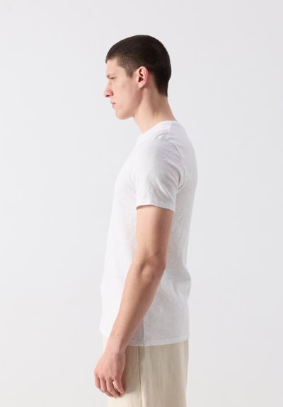 Jeune homme en t-shirt blanc à manches courtes et pantalon beige, debout de profil sur fond blanc.